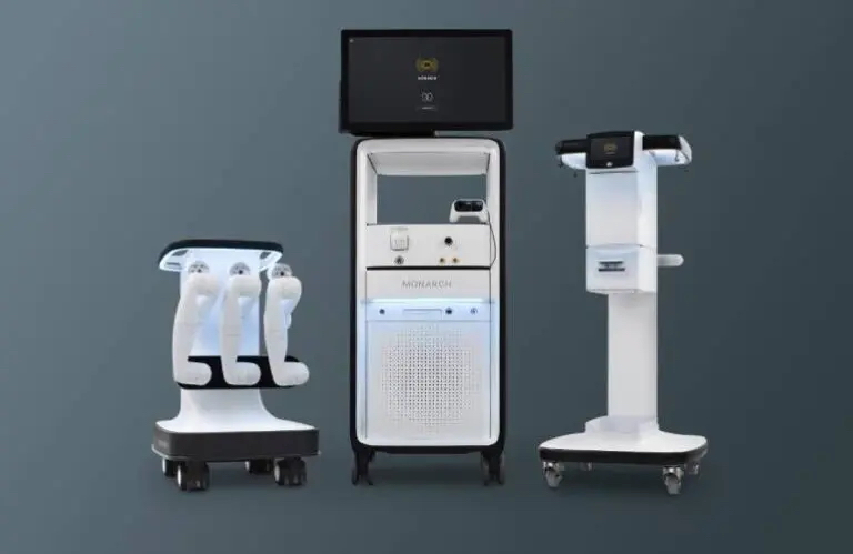 From left to right, a white platform on wheels with three robotic arms, a monitor on a white stand and another white and black stand.