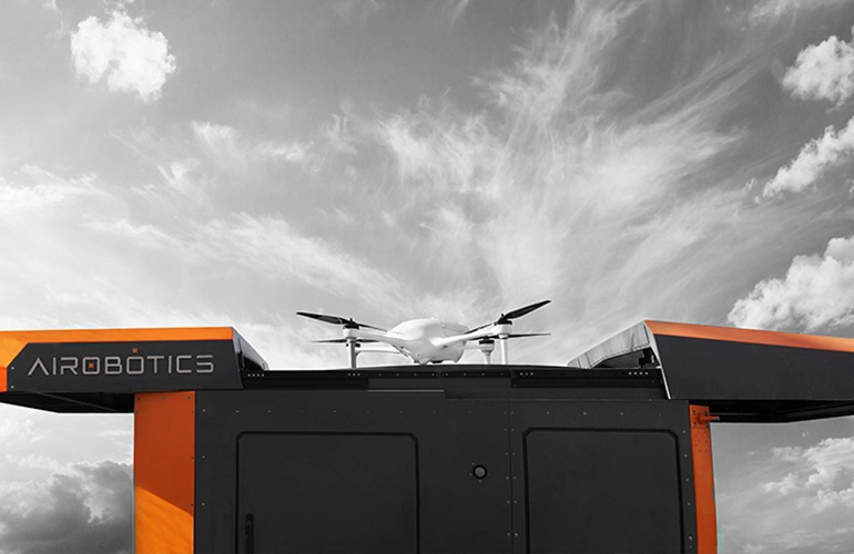 A white drone flying out of a black and grey box labeled "Airobotics" against a black and white sky.