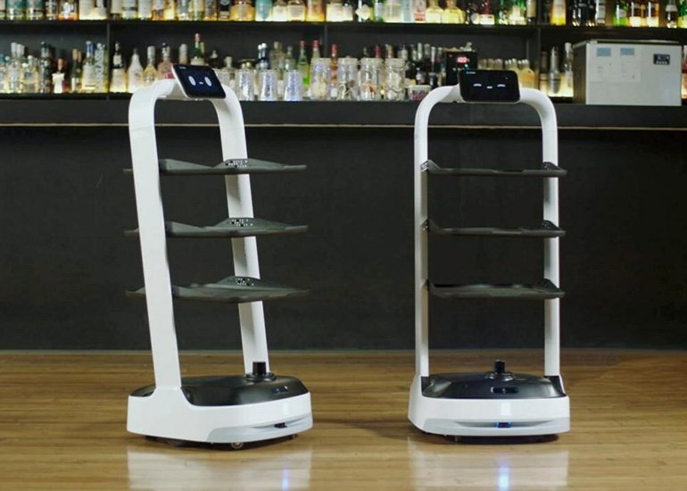 Two white serving robots with three black shelves and a screen with eyes. 