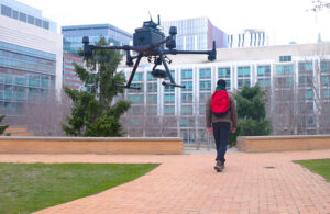 drone flying behind a student with a red backpack.