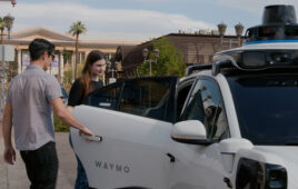 A man opening the door of a waymo vehicle for a woman with palm trees in the background.