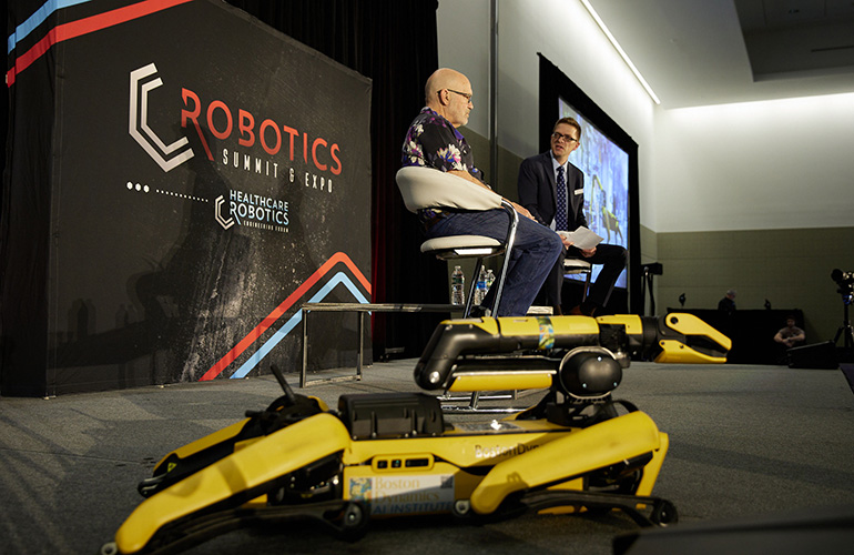 marc raibert and steve crowe on stage at robotics summit 2023 with a boston dynamics spot in the foreground.