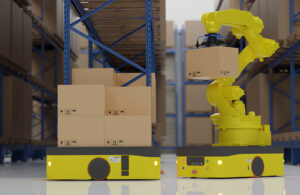 A mobile manipulator robot picking up a box to put on a mobile robot in a warehouse. Behind the robots are racks full of cardboard boxes.