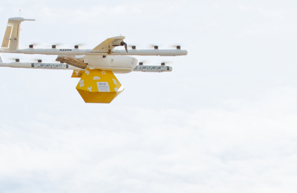 „Wing Aviation“ dronas per dangų neša geltoną paketą.