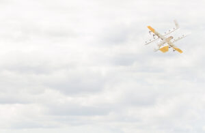 A yellow and white Wing drone in the upper right corner, flying into a cloudy sky.