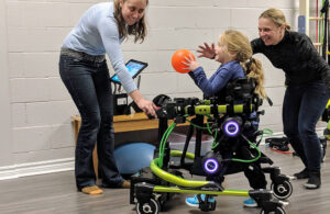 A young girl with long blonde hair using the Trexo Robotics brace to walk and play with a basketball.