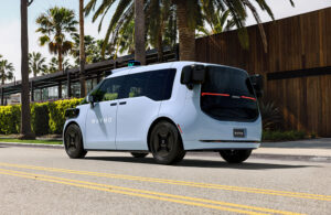 Waymo's 6th generation robotaxi, a boxy light blue vehicle that says "Waymo" on the side, driving on a road lined with palm trees.
