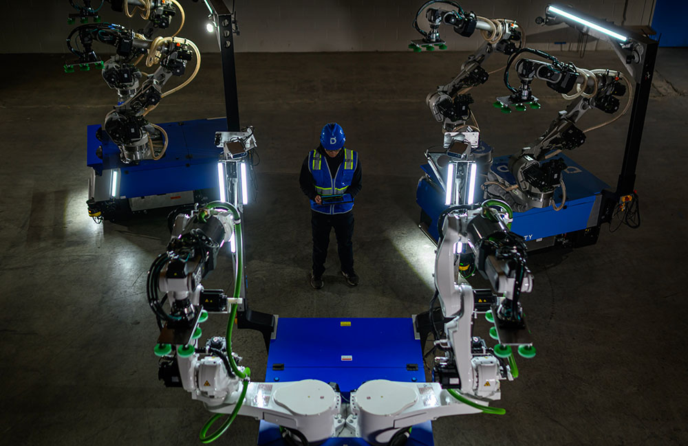 Dexterity's Mech two armed robot in front of a worker in a blue hardhat. The company is working with Sanmina on manufacturing.