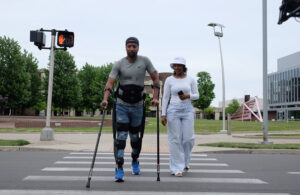 A man walking down a crosswalk wearing the Ekso personal exoskeleton with a woman walking beside him. The man is also using crutches to stay steady.