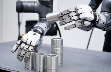 two humanoid robotics hands picking cylindrical items from tables.